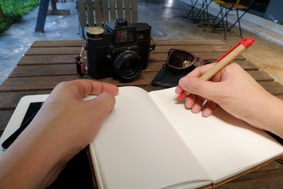Hands writing in a notebook at an outdoor workspace with a camera and sunglasses.