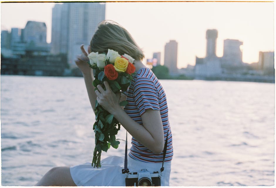 A young woman holds a bouquet of roses near a city waterfront at sunset.